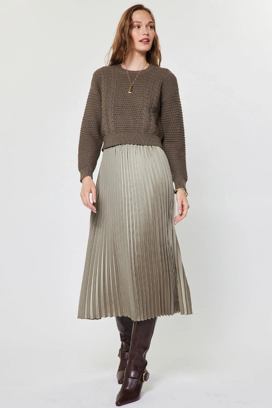 A woman modeling a brown textured knit sweater and a pleated taupe skirt, paired with dark brown leather boots, against a plain light gray studio background.