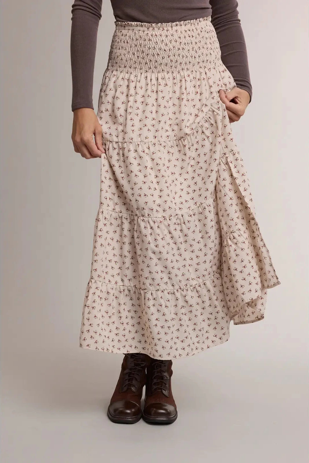 A model wearing a high-waisted, tiered maxi skirt with a small floral print in beige and brown, styled with a dark brown long-sleeve top and dark brown lace-up boots against a neutral studio background.