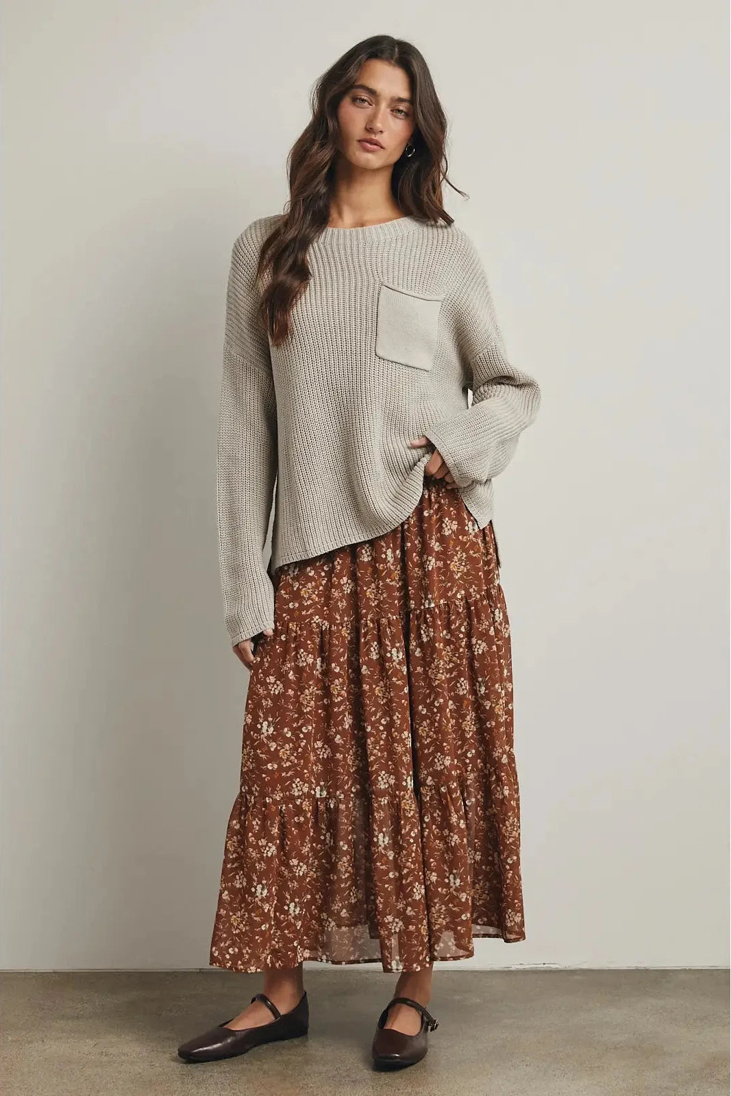 Woman wearing a light gray sweater and brown floral skirt against a plain background