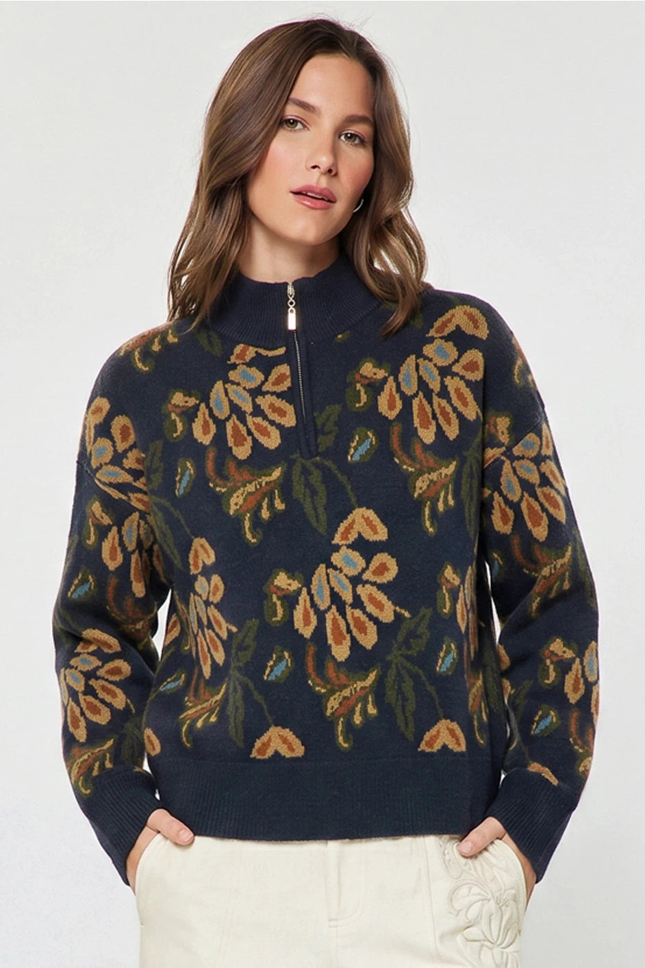 A woman wearing a navy blue quarter-zip sweater with a brown and green floral pattern, paired with off-white pants, against a light gray studio background.