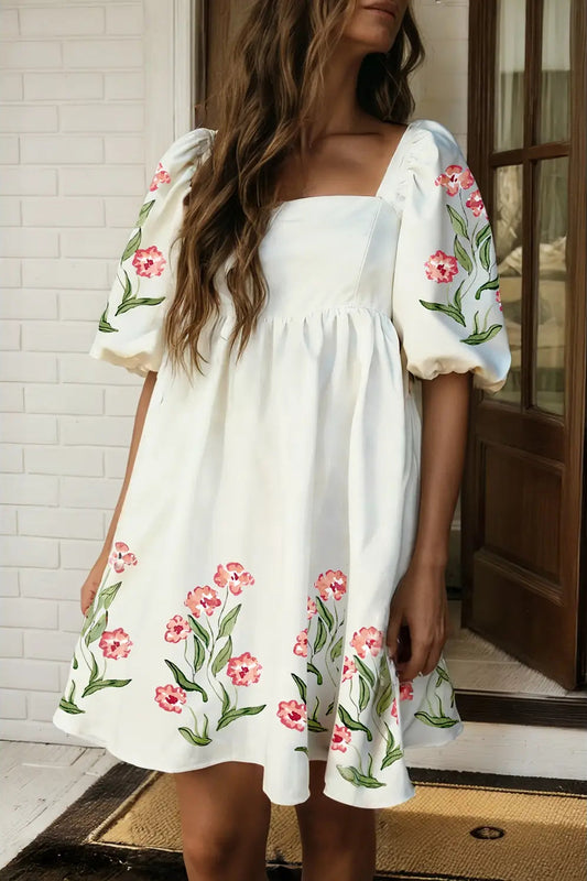 Woman wearing a white dress with floral embroidery standing indoors.