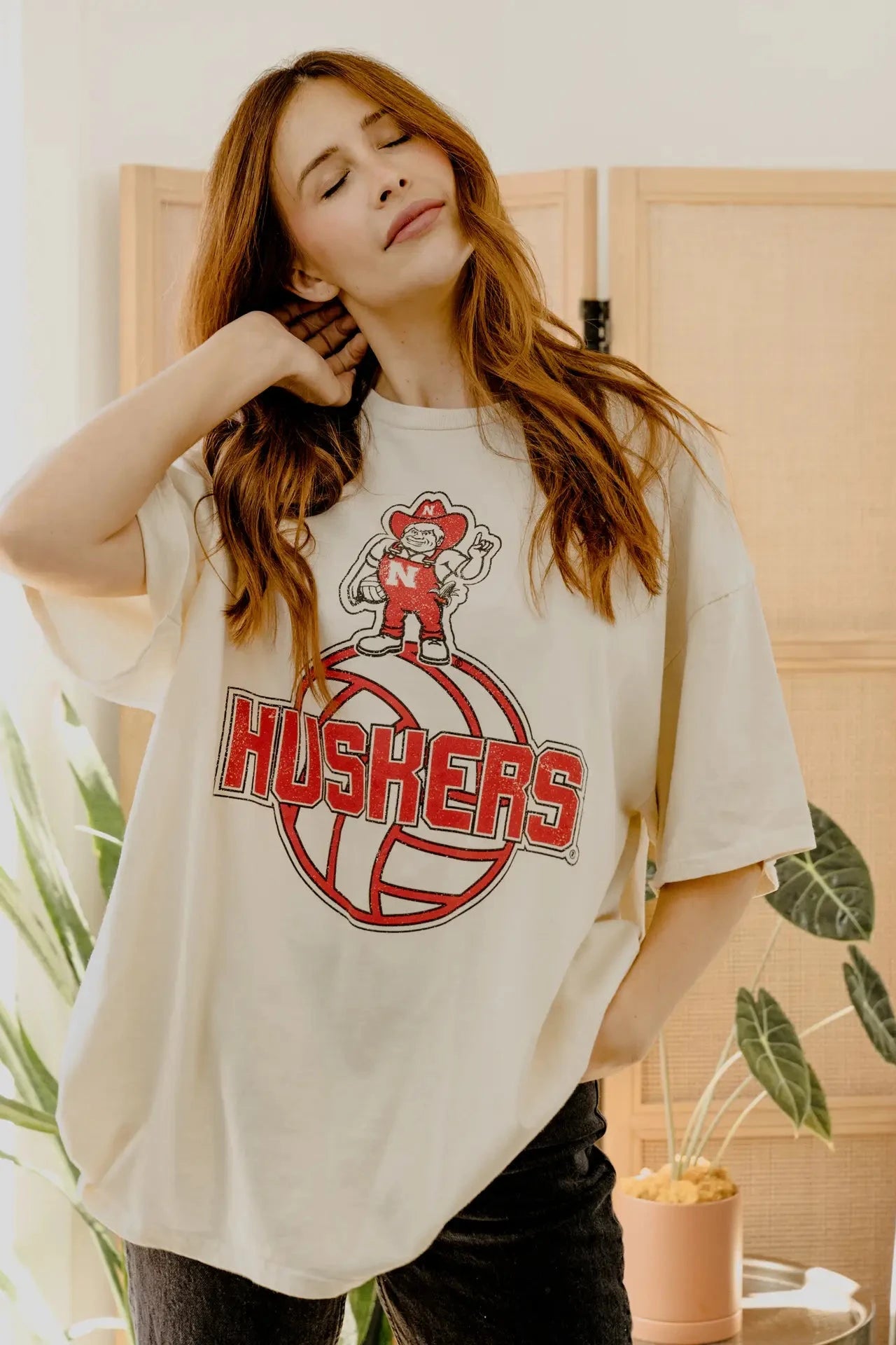 A woman wearing an oversized cream-colored t-shirt with a red and brown 'HUSKERS' mascot graphic, posed in a brightly lit indoor setting with plants and wooden screens.