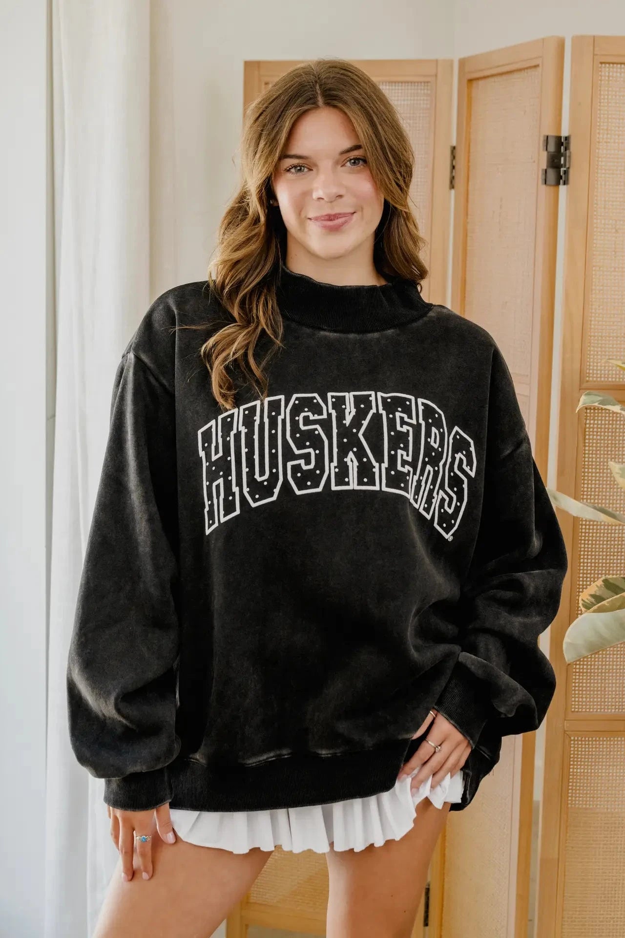 A woman wearing a black distressed sweatshirt with 'HUSKERS' printed across the chest in white varsity-style lettering, standing indoors in front of a light wood folding screen.