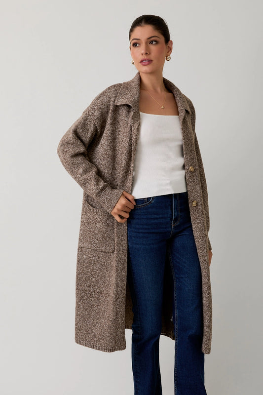 Woman wearing a long brown coat over a white top and blue jeans on a plain background