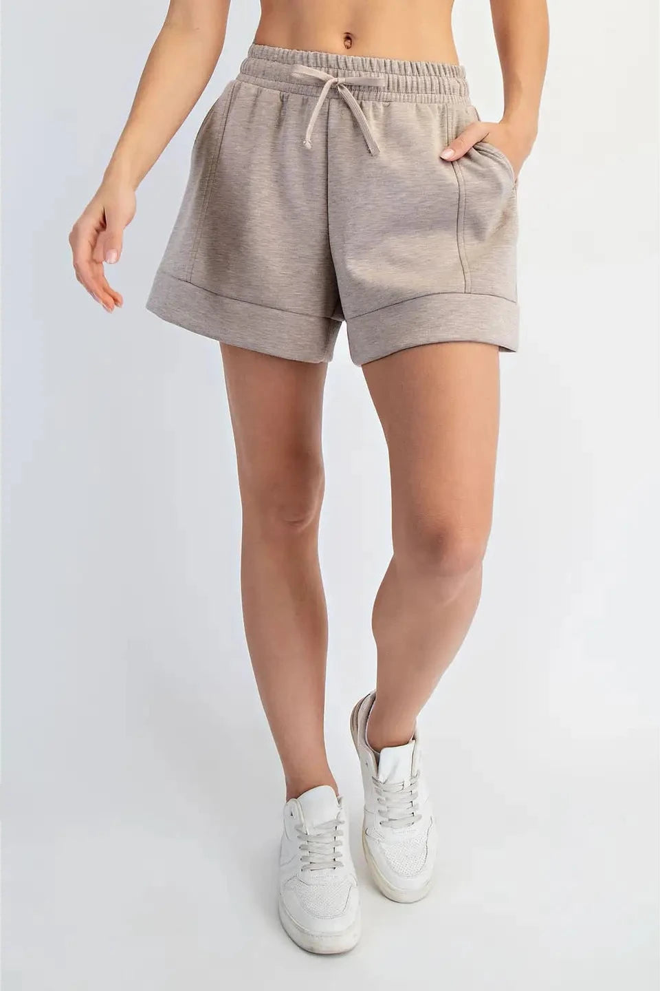 A model wearing light gray heathered sweat shorts with a drawstring waist and rolled cuffs, paired with white sneakers, shown from the waist down against a light gray studio background.