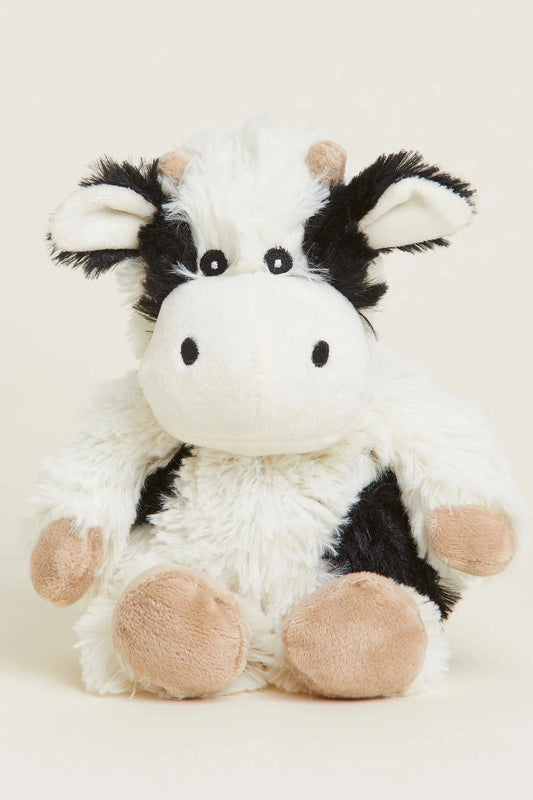 A plush cow toy with a fluffy white body, black patches, and beige hooves and ears, sitting against a plain light beige background.