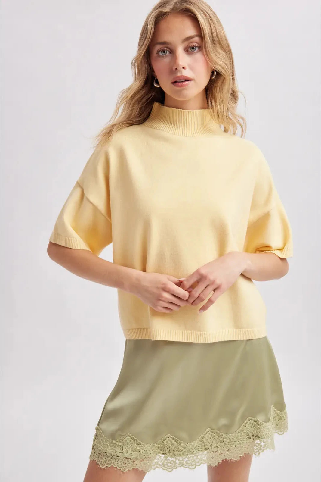 A woman modeling a pale yellow short-sleeve mock neck knit sweater paired with an olive green satin mini skirt featuring a lace hem, set against a plain light gray studio background.