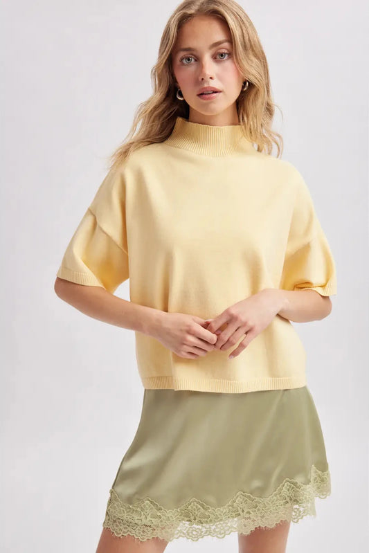 A woman modeling a pale yellow short-sleeve mock neck knit sweater paired with an olive green satin mini skirt featuring a lace hem, set against a plain light gray studio background.