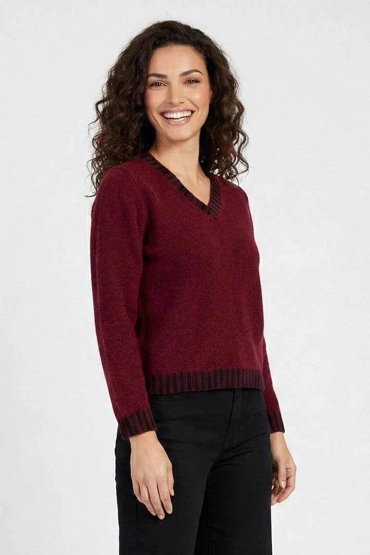 A woman with curly dark hair smiling, wearing a maroon knit V-neck sweater with dark trim and black pants, photographed against a light gray studio background.