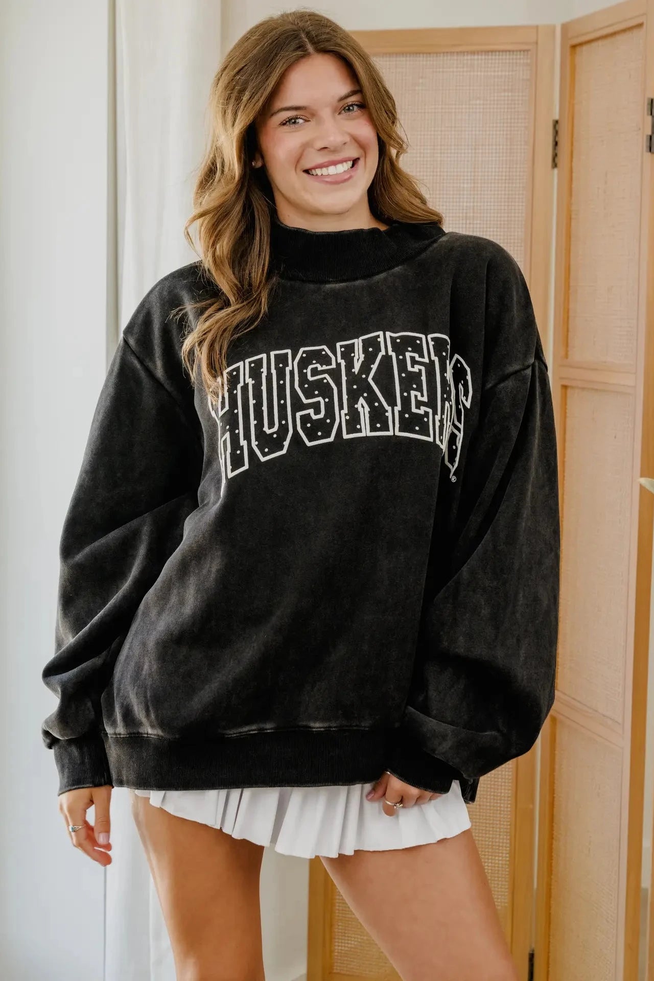 A woman wearing an oversized black washed sweatshirt with 'HUSKERS' printed across the chest in white outlined block letters, paired with a white pleated skirt, standing indoors in front of a light wood screen door.