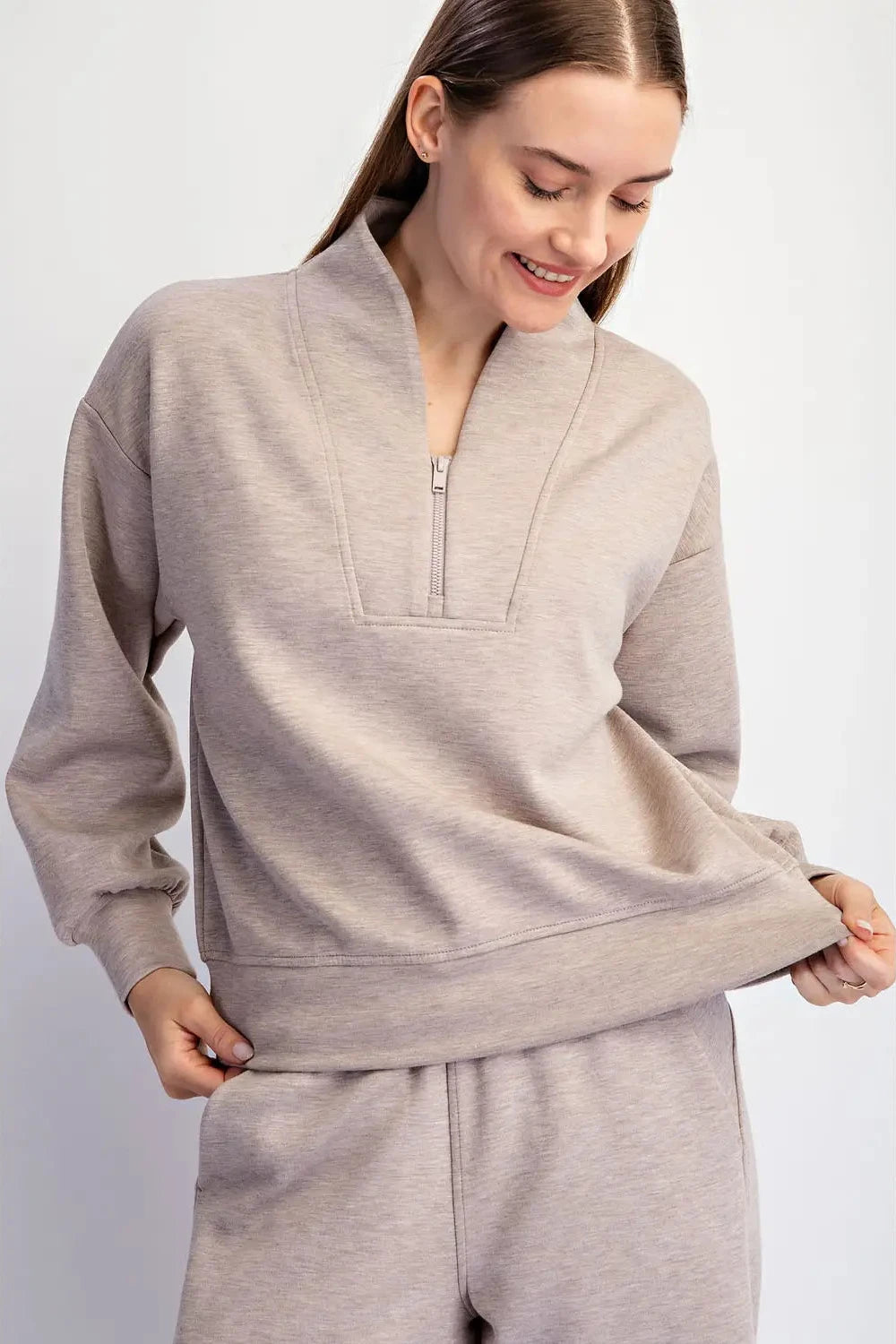 A woman wearing a light heather gray half-zip sweatshirt and matching sweatpants, shown from the chest down against a plain light gray studio background.