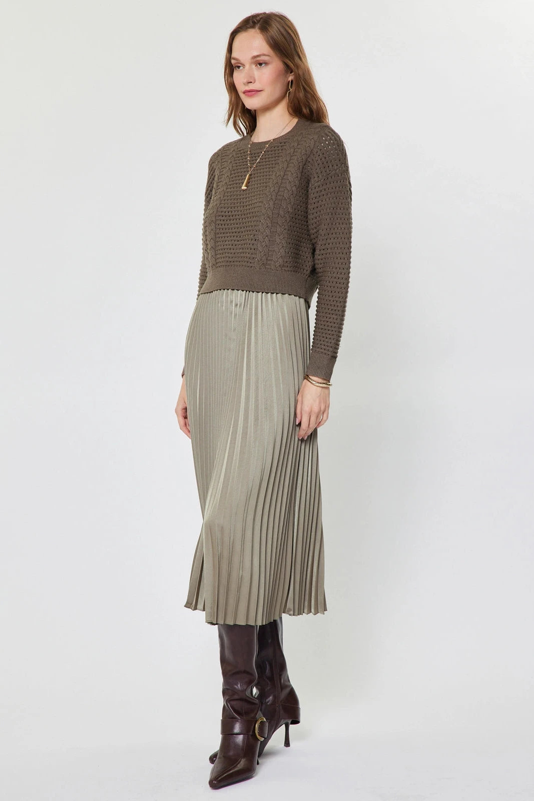 A woman modeling a brown textured knit long-sleeve sweater paired with a pleated light beige midi skirt and dark brown leather ankle boots, shot against a plain light gray studio background.