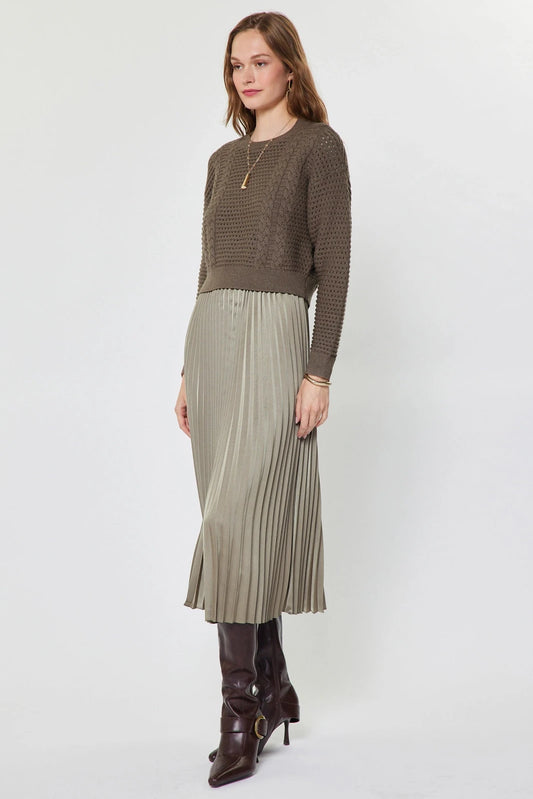 A woman modeling a brown textured knit long-sleeve sweater paired with a pleated light beige midi skirt and dark brown leather ankle boots, shot against a plain light gray studio background.