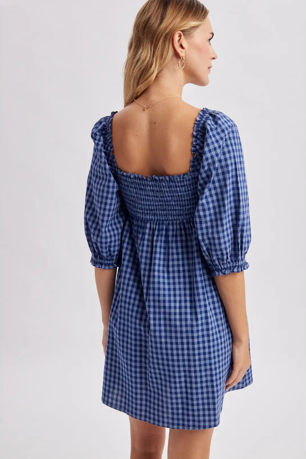 A woman wearing a blue and white gingham mini dress with puff sleeves and a smocked back neckline, shown from a rear three-quarter view against a light gray studio background.