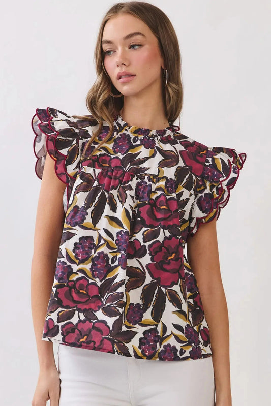 A woman wearing a white blouse with a bold floral print in dark purple, magenta, and olive green, featuring ruffled cap sleeves, paired with white pants against a light gray studio background.
