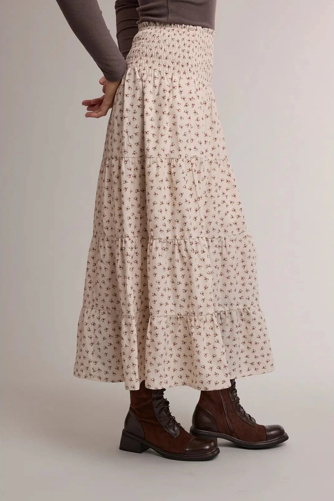 A model is shown from the waist down wearing a cream-colored, tiered maxi skirt with a small brown floral pattern, paired with dark brown lace-up boots against a neutral studio background.