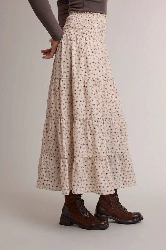 A model is shown from the waist down wearing a cream-colored, tiered maxi skirt with a small brown floral pattern, paired with dark brown lace-up boots against a neutral studio background.