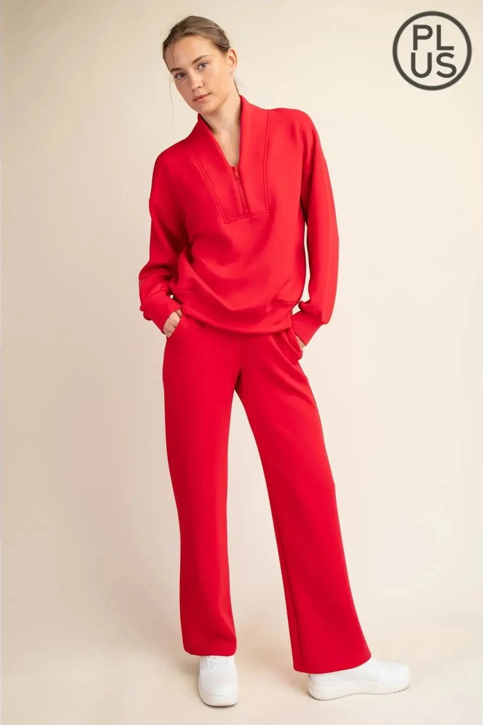 A woman modeling a bright red two-piece tracksuit with a half-zip long-sleeve top and matching wide-leg pants, paired with white sneakers, against a light beige studio background. A circular 'PLUS' logo is visible in the top right corner.