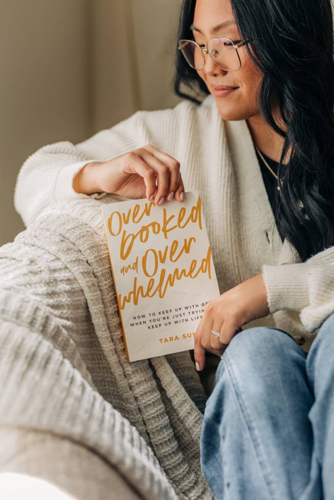 Woman holding a book titled 'Overbooked and Overwhelmed' in a cozy indoor setting.