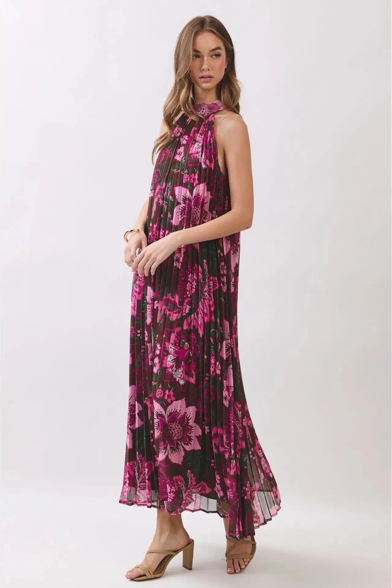 A woman modeling a sleeveless halter-neck maxi dress with a dark background and a vibrant magenta floral print, paired with tan heeled sandals against a light gray studio backdrop.