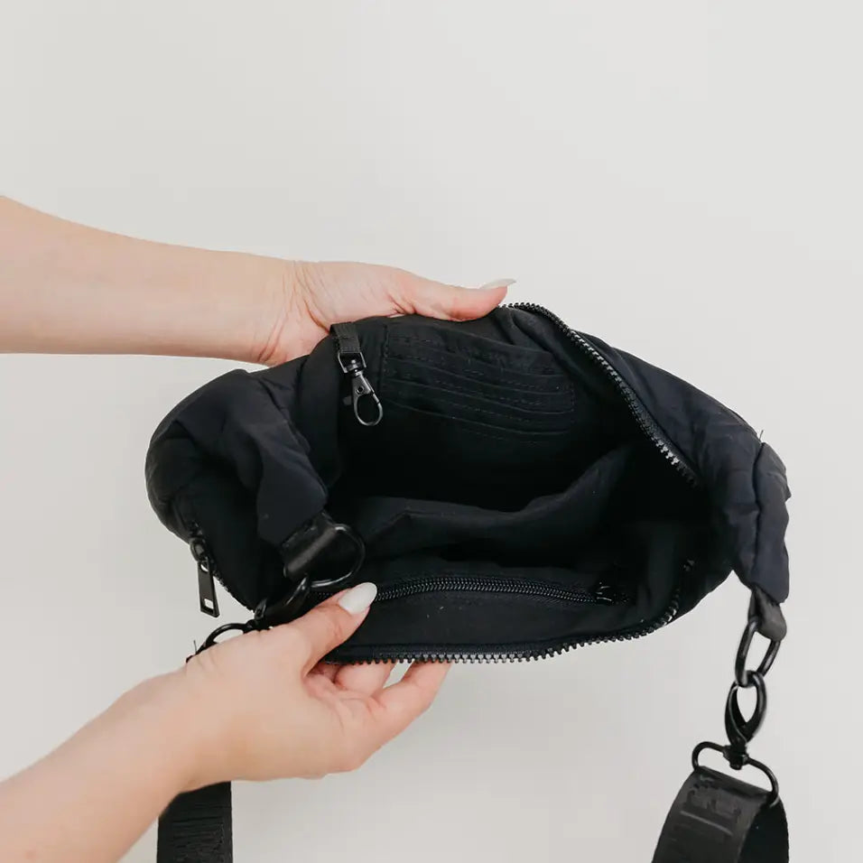 A black nylon crossbody bag is shown open, held by two hands to display its interior compartments, zipper details, and strap attachments against a plain light gray background.