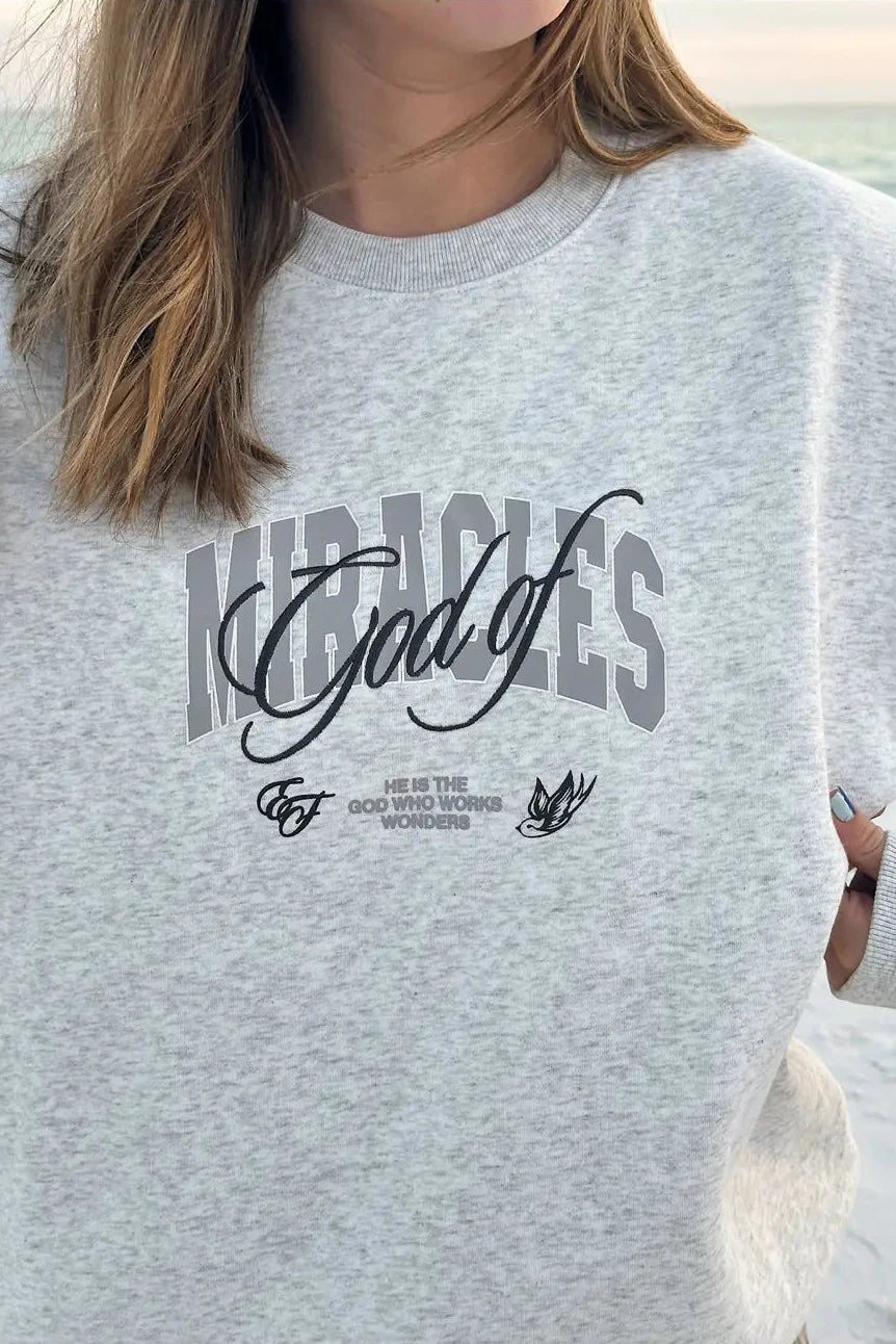 Person wearing a gray sweatshirt with 'Miracles God of' text design.