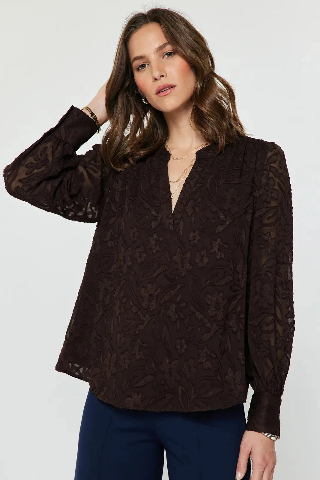 A woman wearing a dark brown long-sleeve lace blouse with a split neckline and navy blue pants, photographed against a light gray studio background.