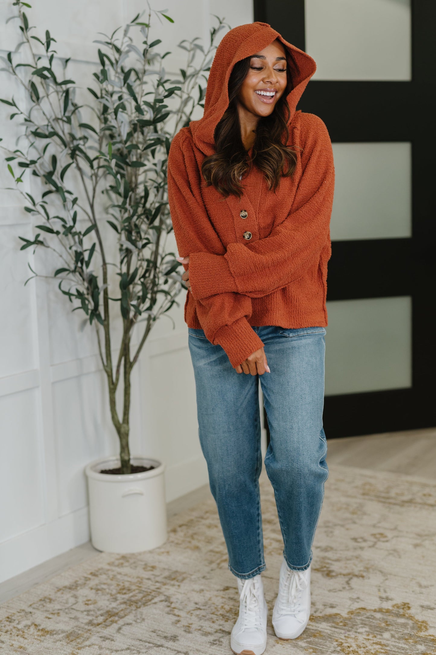 Woman wearing a rust-colored hoodie and blue jeans standing indoors next to a plant.