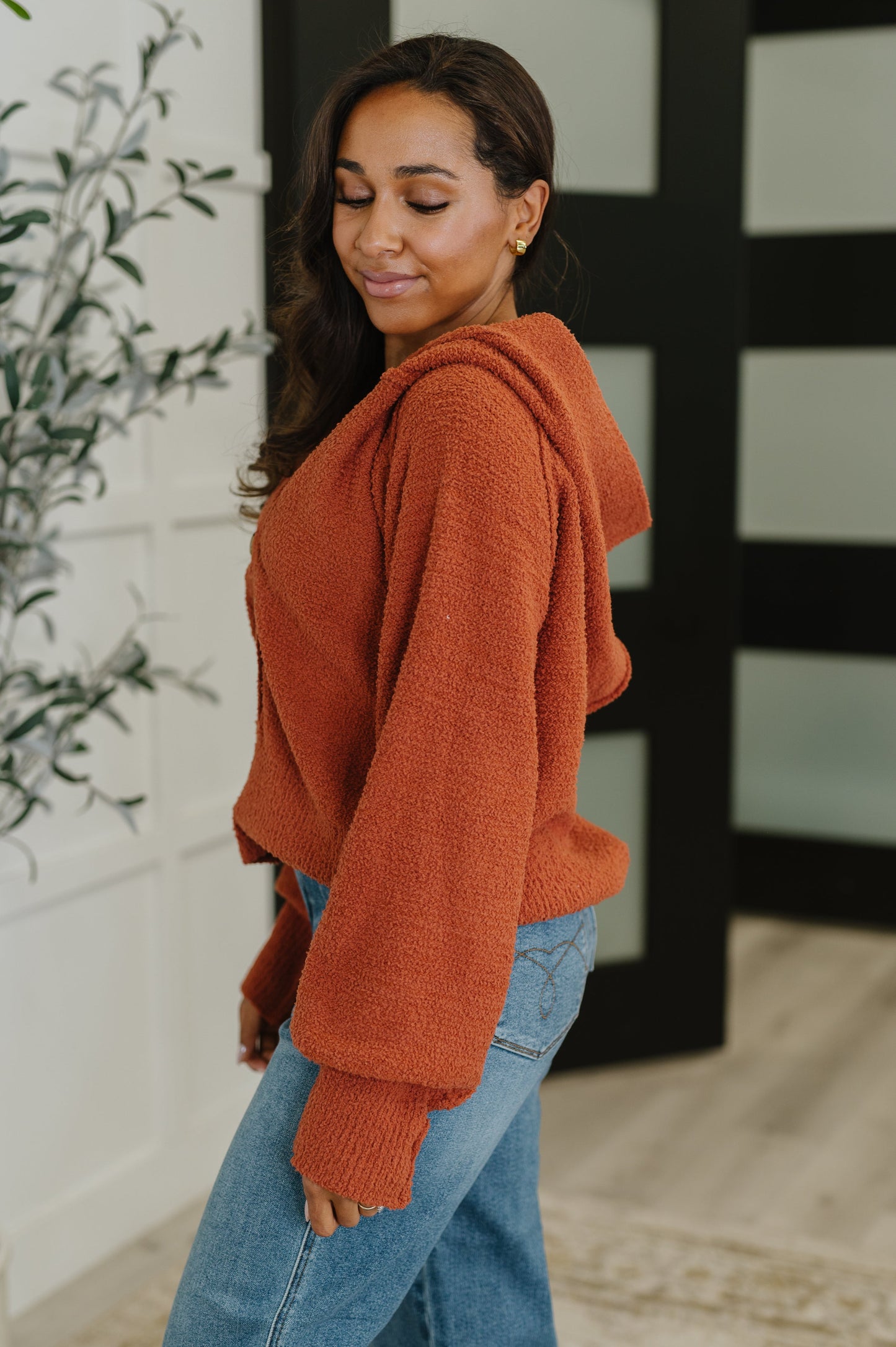 Woman wearing a rust-colored hoodie and blue jeans standing indoors.