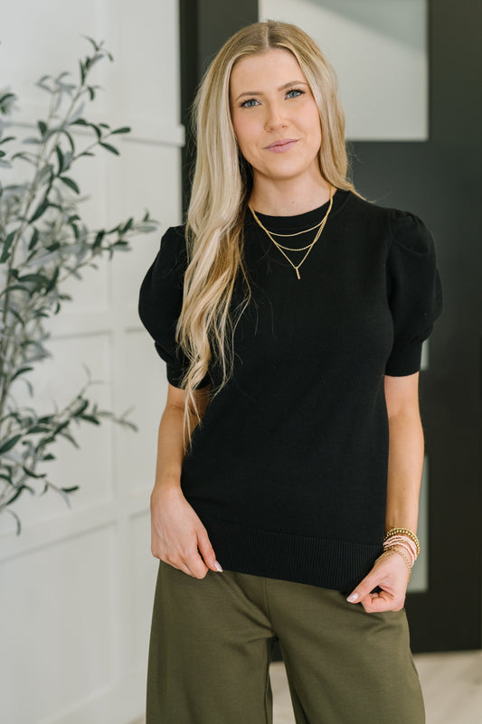 Woman wearing a black top and green pants with a blurred indoor background