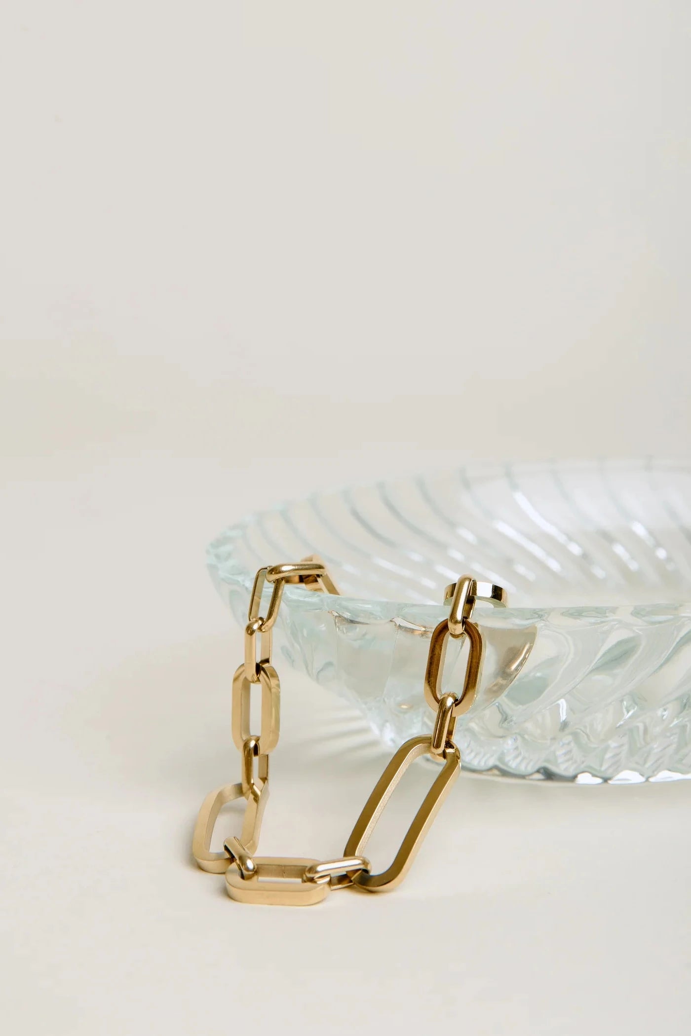 Gold chain bracelet on a textured white surface with a clear glass dish in the background