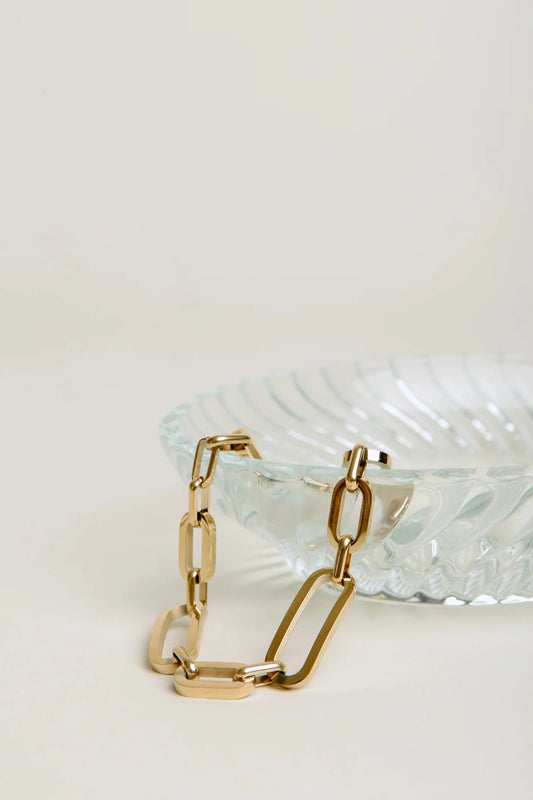 Gold chain bracelet on a textured white surface with a clear glass dish in the background