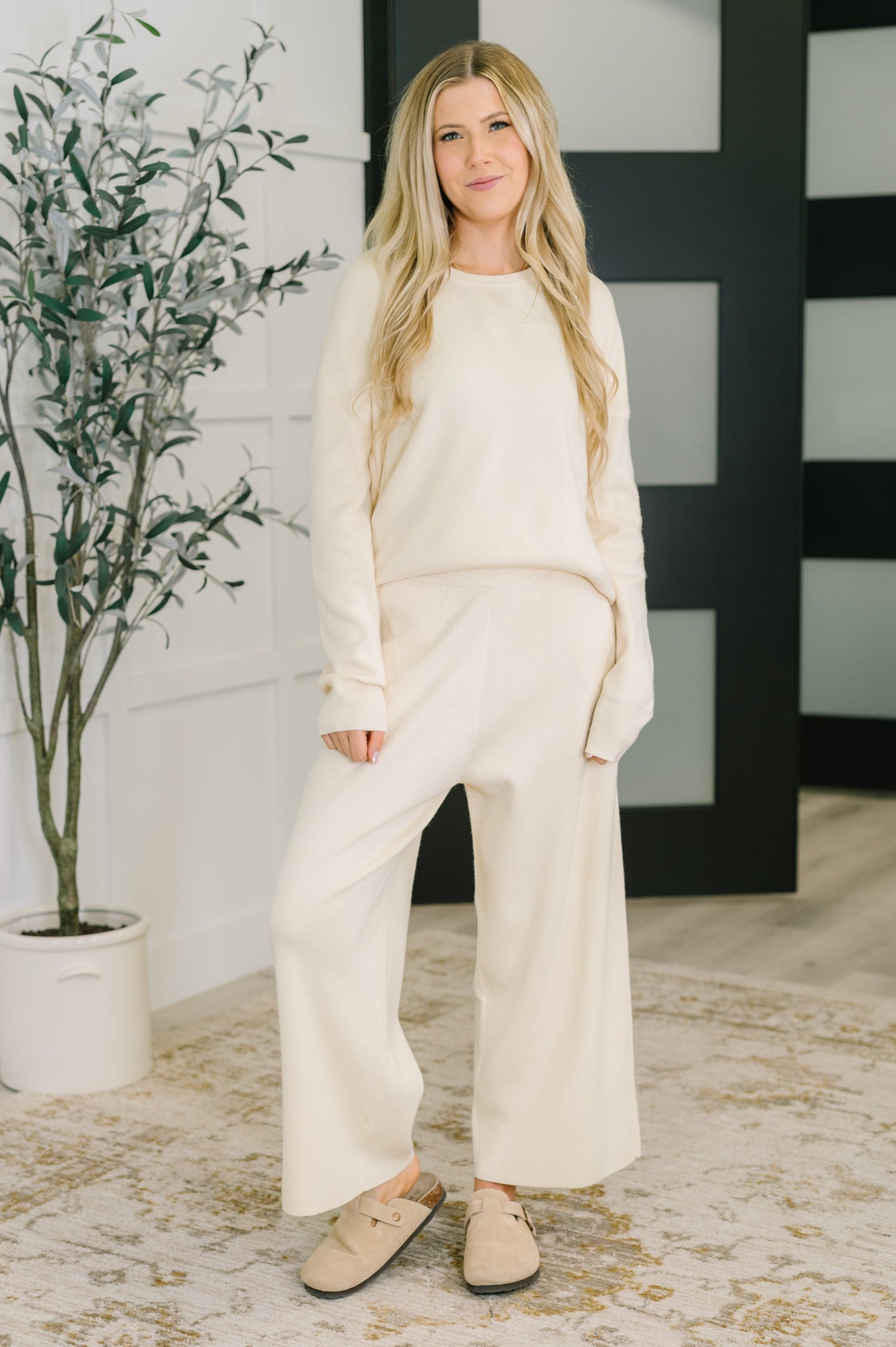 Woman in a cream-colored outfit standing indoors with a plant and decorative wall in the background