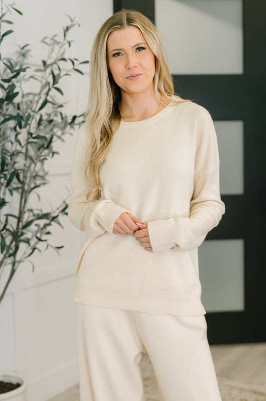 Woman wearing a cream-colored outfit standing indoors with a plant in the background