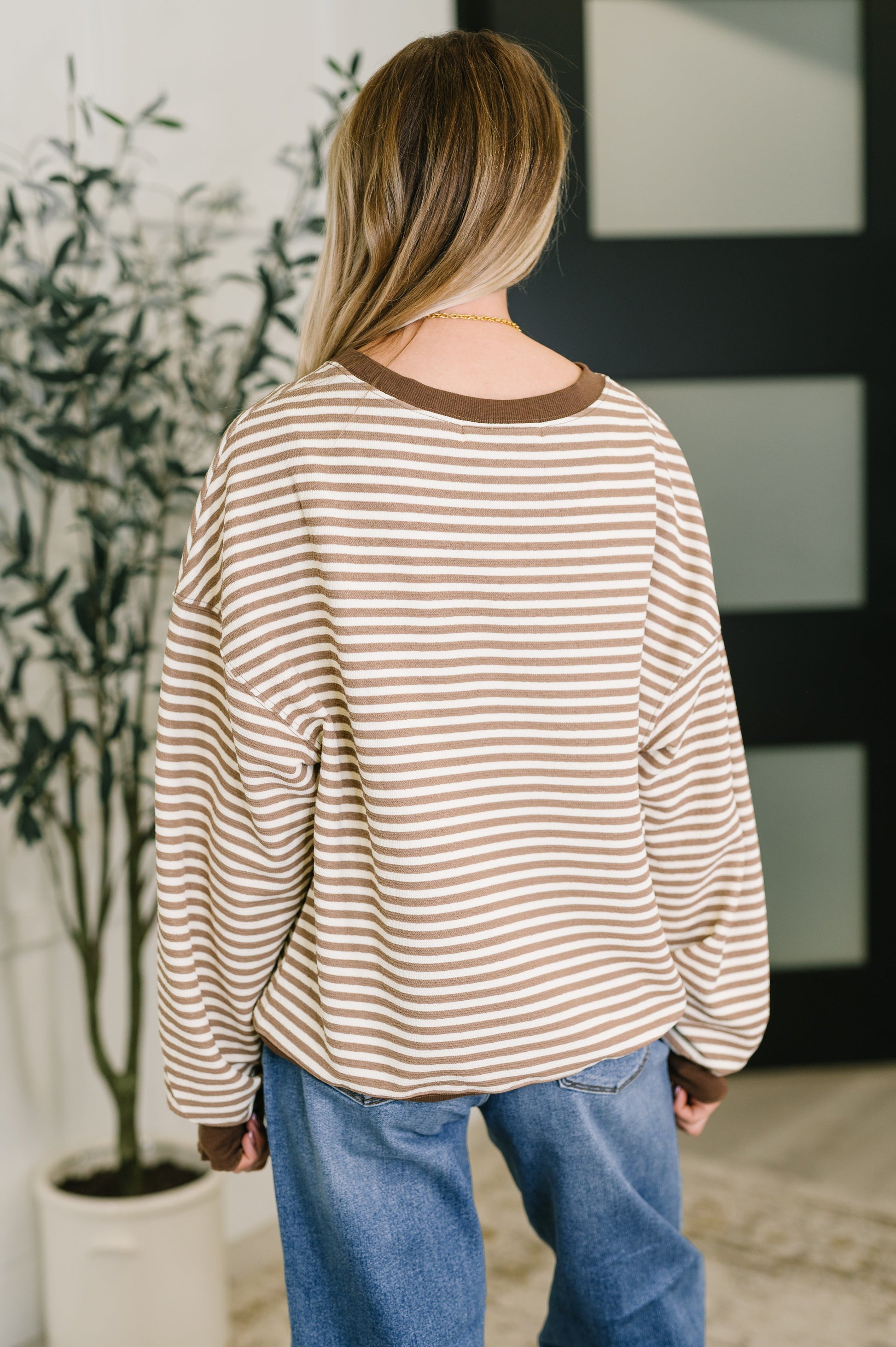 Person wearing a striped shirt and blue jeans standing in a room with a plant and framed picture.