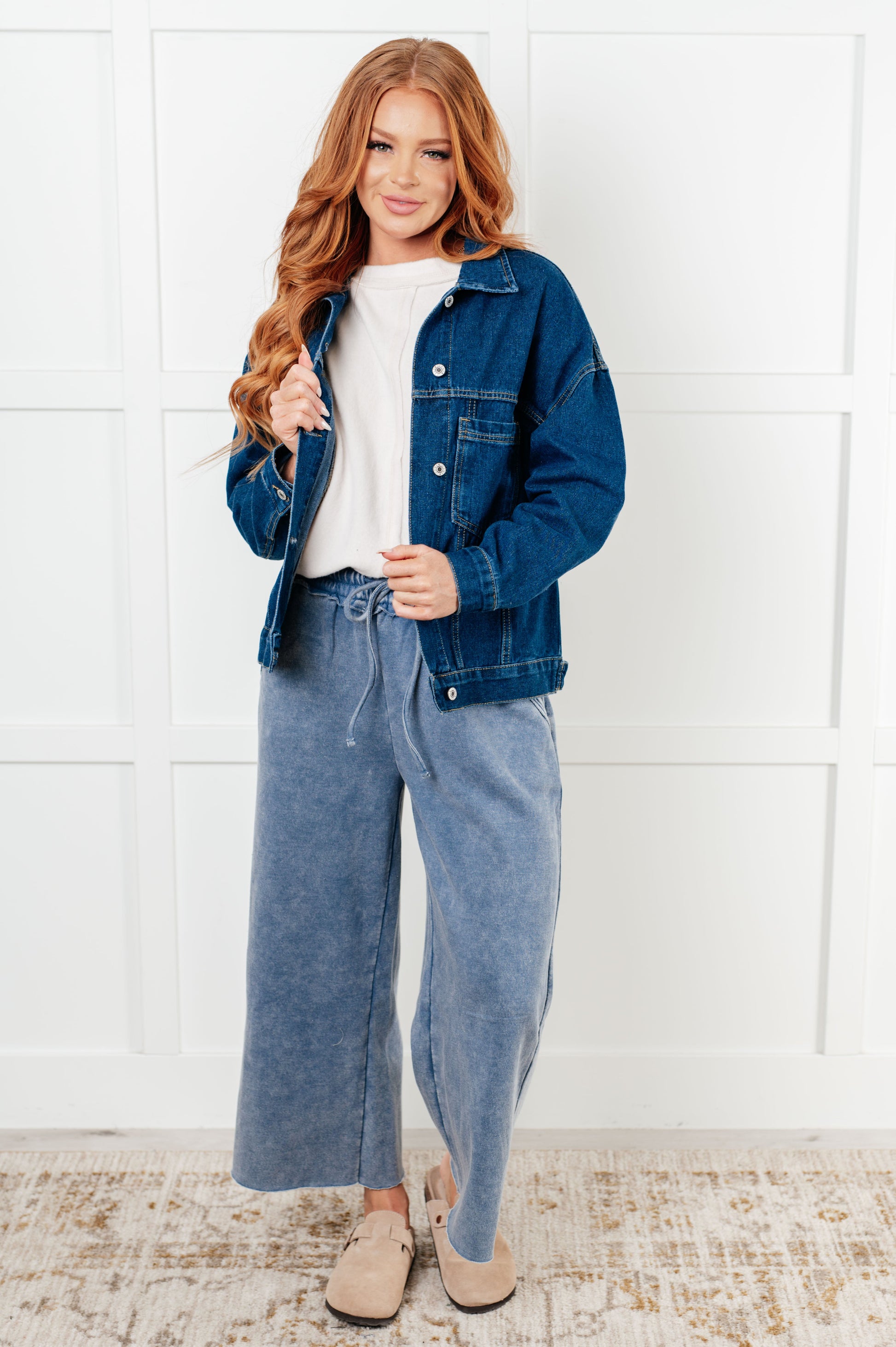 Woman wearing a blue denim jacket and wide-leg jeans against a white wall.