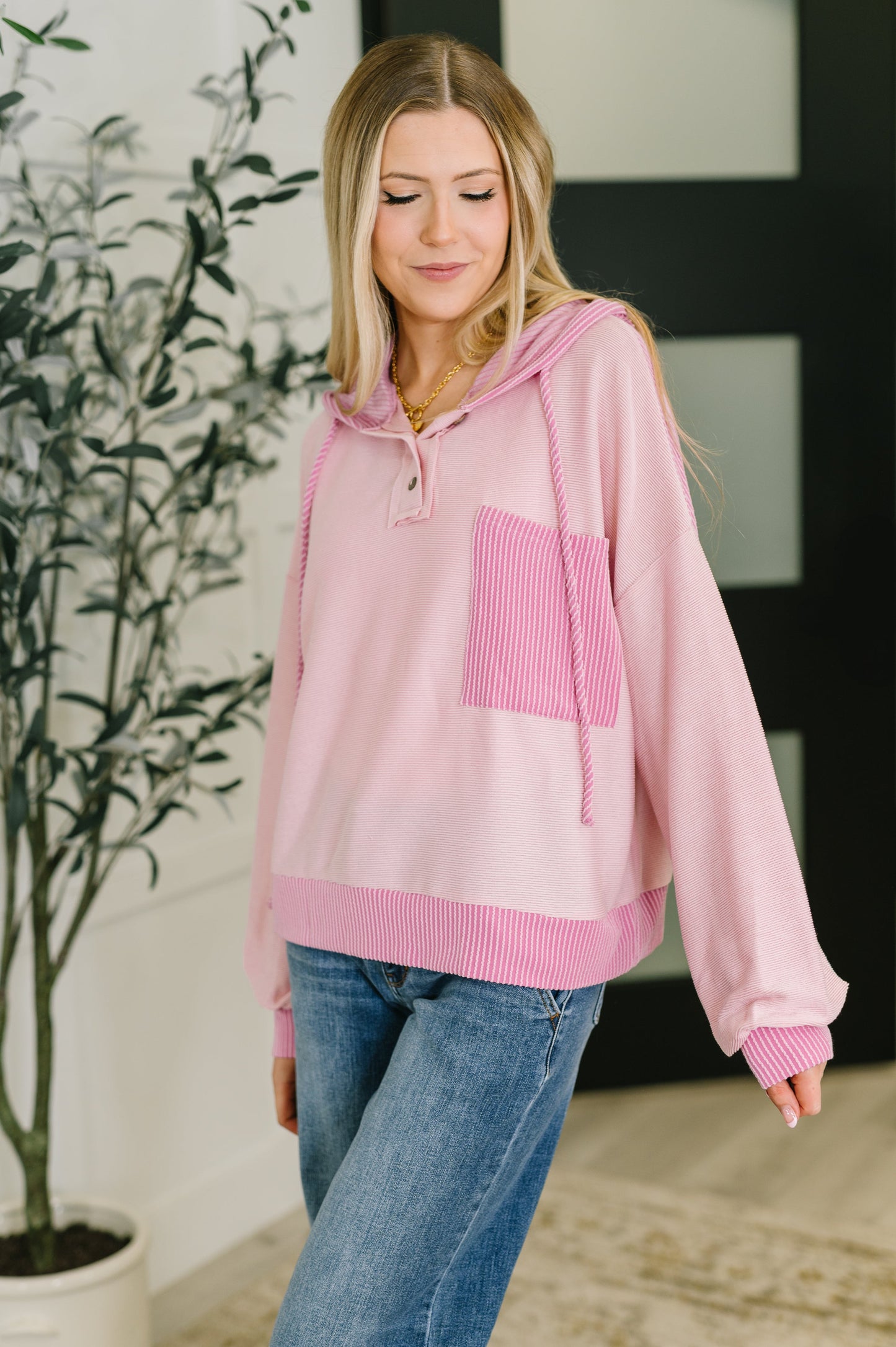 Woman wearing a pink hoodie with a pocket, standing indoors next to a plant.