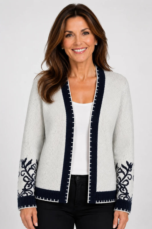 A woman wearing a light gray knit cardigan with dark navy blue trim and matching navy blue sleeve embroidery, styled over a white top and black pants, against a light gray studio background.
