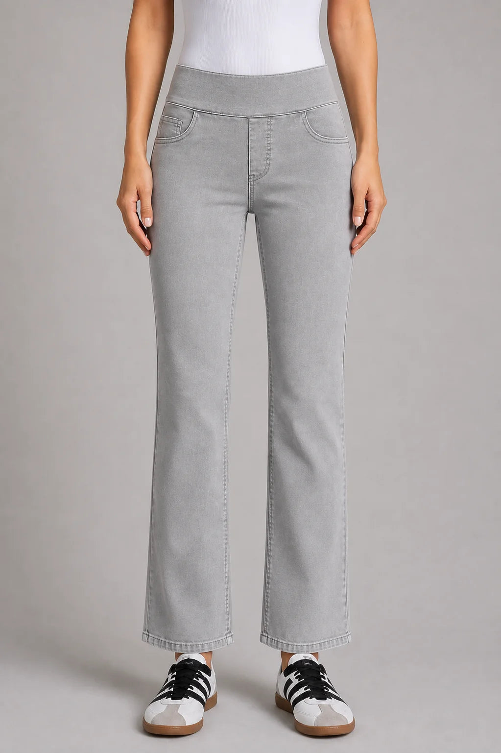 A model wearing light gray wide-leg jeans with a straight-to-slightly-flared leg, paired with a white top and white sneakers with black stripes, against a solid gray studio background.