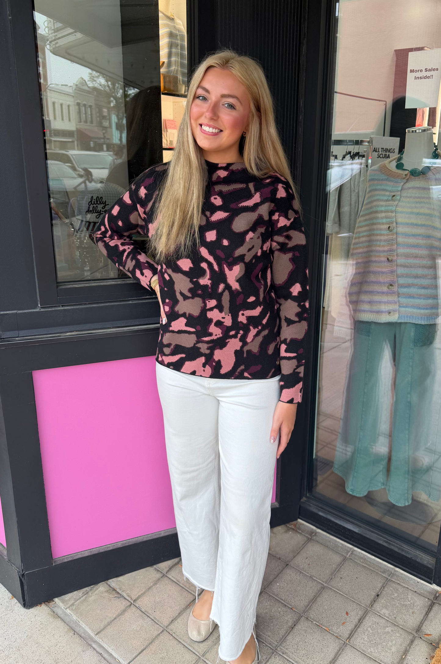 A woman stands in front of a boutique storefront wearing a sheer black mock-neck top with a pink abstract camouflage pattern and white wide-leg pants.