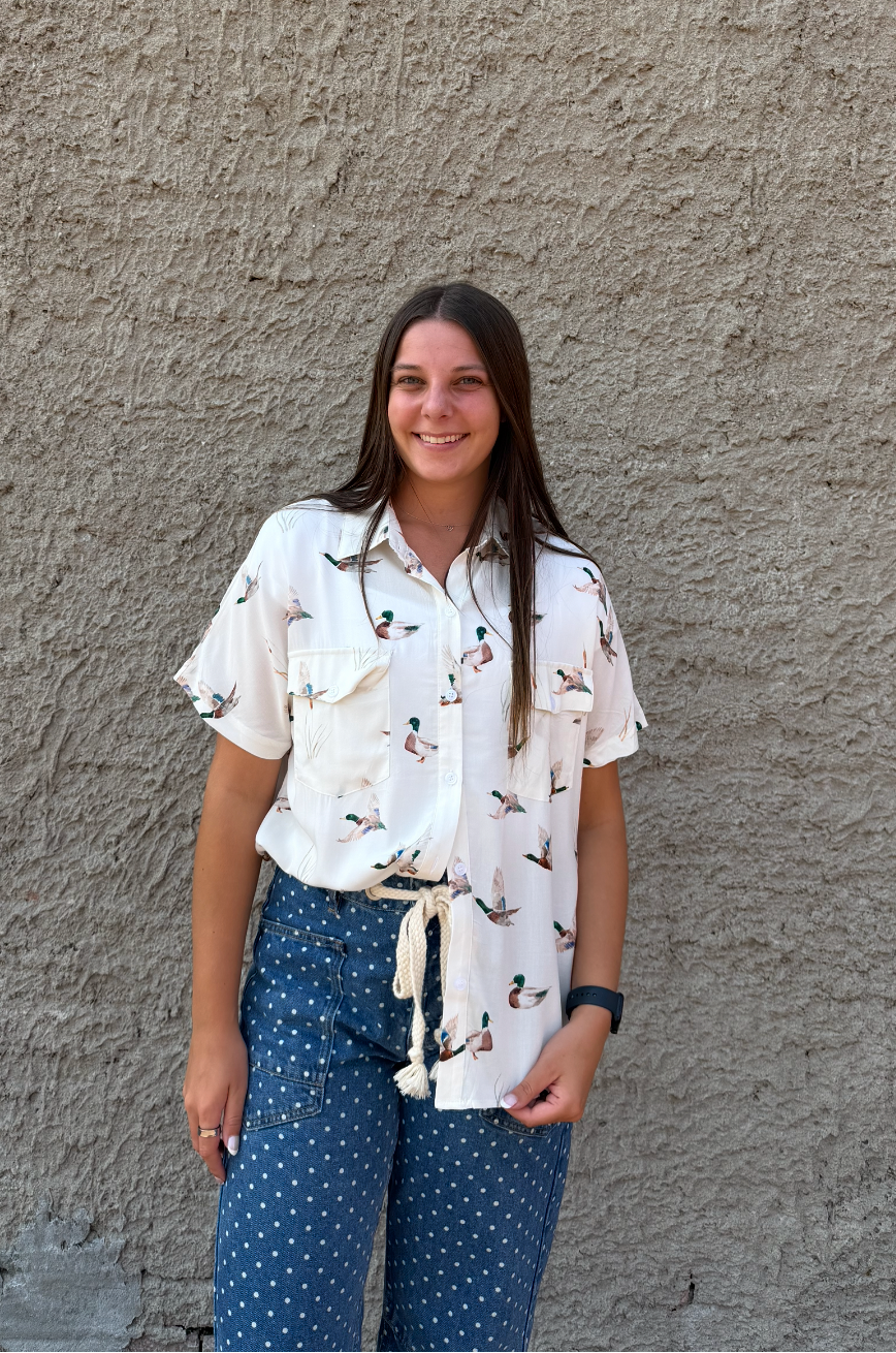 A woman stands against a textured gray stucco wall wearing a white short-sleeve button-up shirt with a small black pattern and blue patterned pants.
