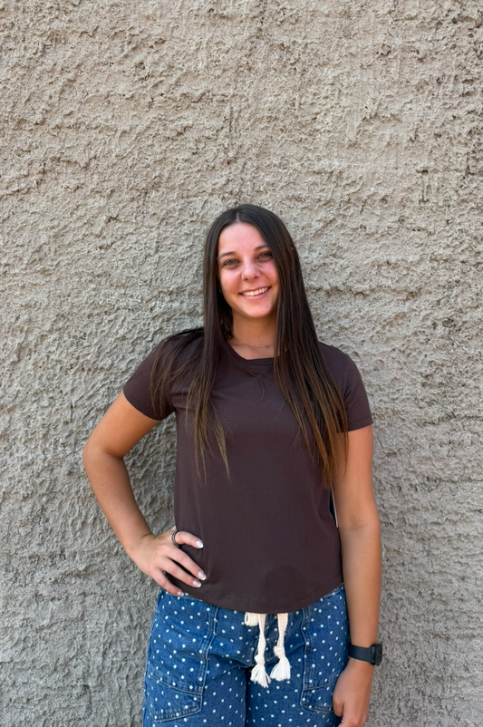 A woman with long dark hair stands against a textured concrete wall, wearing a dark brown short-sleeve t-shirt and blue patterned shorts with a white drawstring.
