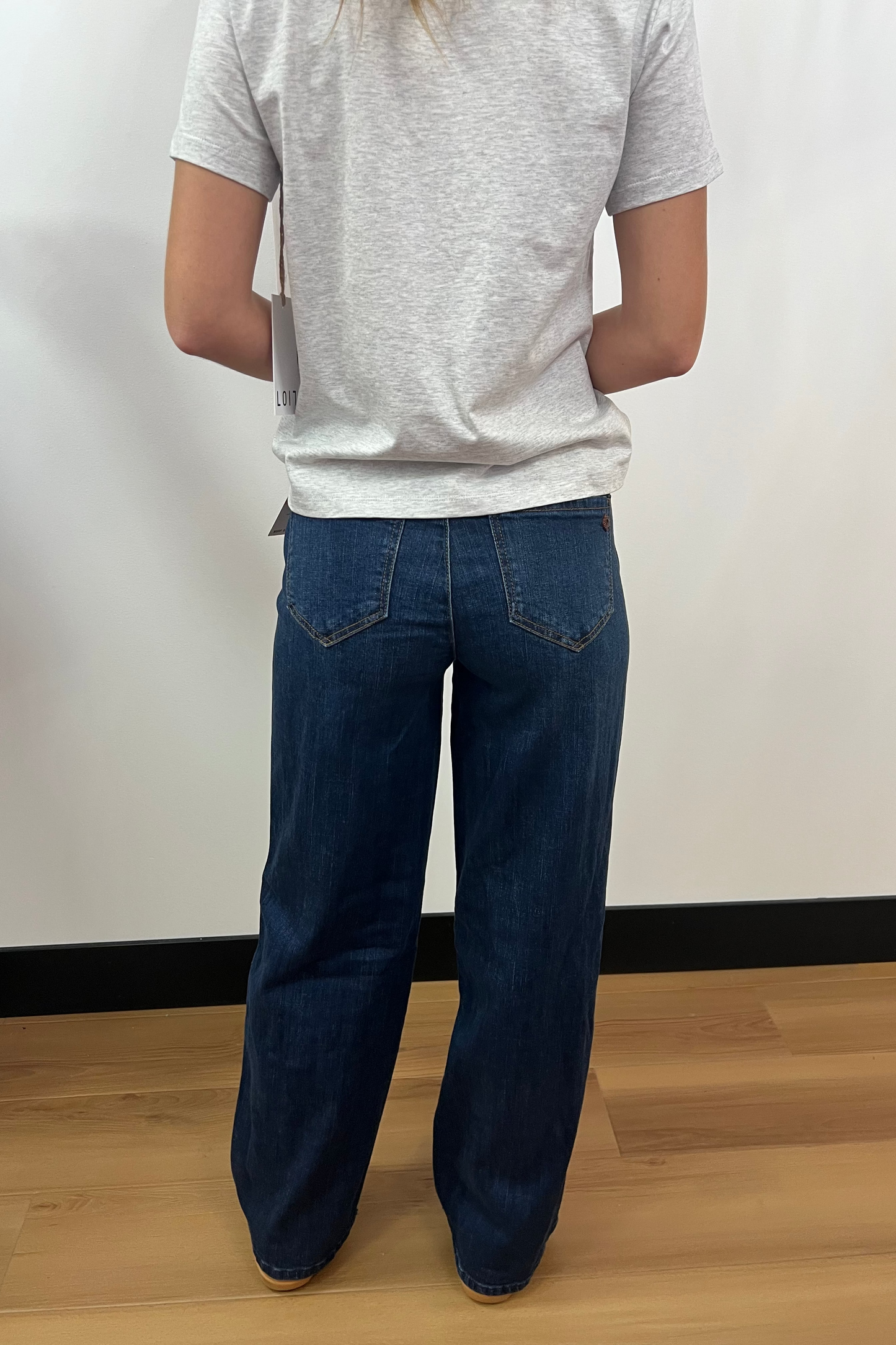 Person wearing a gray t-shirt and blue jeans standing against a white wall.