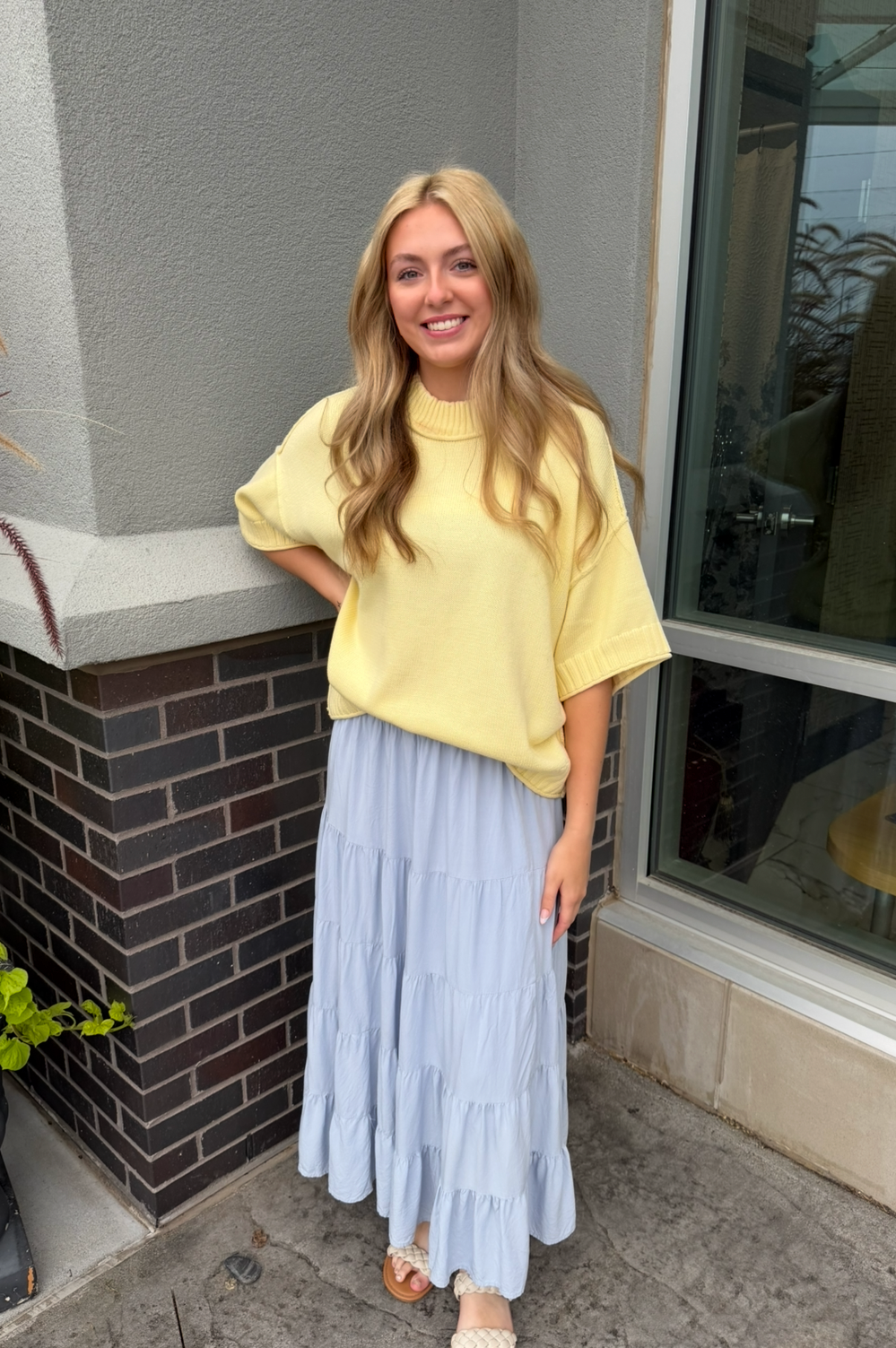 A woman stands outdoors wearing a loose short-sleeve yellow top and a long light blue tiered skirt, with beige sandals. The photo is taken in front of a building with a gray wall, brick base, and large window.