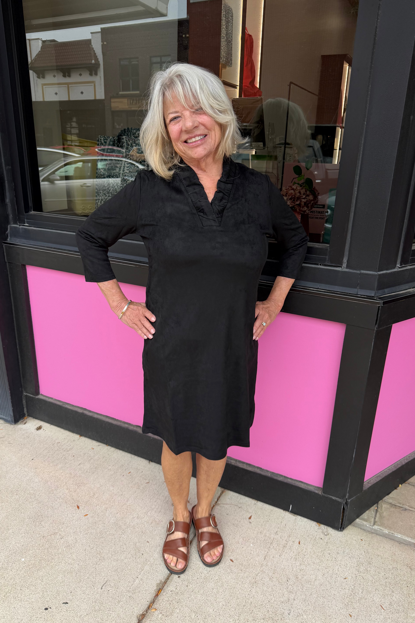 A smiling woman with short gray hair stands outdoors in front of a storefront window, wearing a black knee-length dress with three-quarter sleeves and brown strappy sandals. The building has bright pink panels and a black frame.
