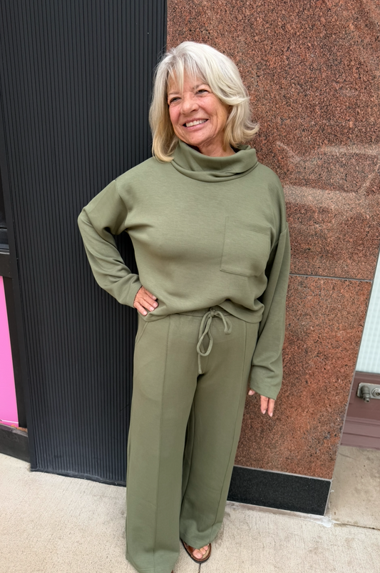 A woman with blonde hair wearing an olive green two-piece lounge set consisting of a mock neck long-sleeve top and matching wide-leg pants, standing outdoors in front of a building wall.