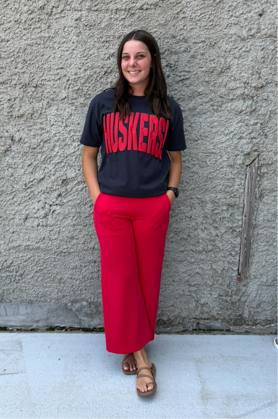 A woman stands against a textured concrete wall wearing a dark gray t-shirt with 'MUSKETEERS' printed in red, paired with bright red wide-leg pants and brown sandals.