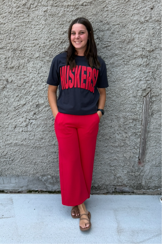 A woman stands against a textured concrete wall wearing a dark gray t-shirt with 'MUSKETEERS' printed in red, paired with bright red wide-leg pants and brown sandals.
