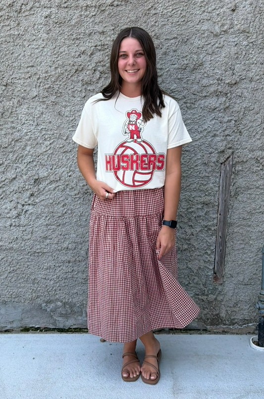 A woman stands outdoors against a textured concrete wall wearing a cream graphic t-shirt with a red 'HUSKERS' logo and a dusty rose pleated midi skirt.