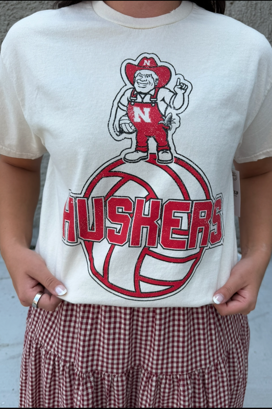 A person wearing a cream-colored vintage short-sleeve t-shirt with a red and white Nebraska Huskers volleyball mascot graphic, paired with a red and white gingham skirt.
